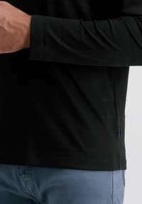 Schwarzes Langarmshirt mit dezenten horizontalen Streifen, rundem Halsausschnitt und Etiketten am Saum, kombiniert mit hellblauen Hosen.