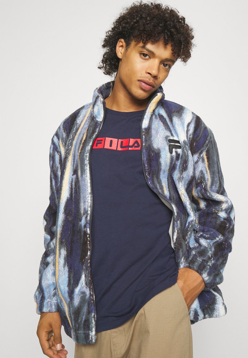 Giacca in pile con un motivo astratto multicolore blu, nero e giallo. Chiusura con zip, colletto rialzato e polsini a coste. Indossata sopra una maglietta blu navy.