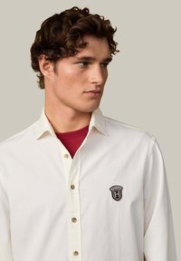 Camisa blanca de botones con cuello, presenta botones marrones y un logo bordado en el lado izquierdo del pecho. El fondo es neutro.