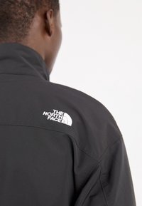 Chaqueta negra hecha de un material liso, con un cuello alto y un logo blanco en el hombro; diseño sencillo.