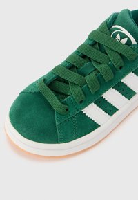 Grønne mokkasinsneakers med hvite lærstriper, med teksturerte grønne lisser og en gummisåle med lyse oransje aksenter.