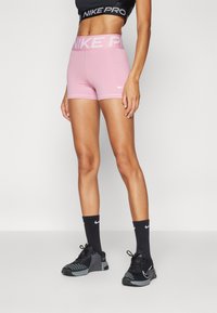 Rózsaszín Nike Pro rövidnadrág sz�éles elasztikus derékpánttal, amelyen ismétlődő fehér "NIKE PRO" felirat található. Fekete Nike zoknik és sportcipők szürke részletekkel.