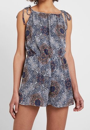 Kvinde iført en ærmeløs, mønstret romper med skulderbånd og elastisk talje, med cirkulære og blomsterdesign i blå og brune nuancer.