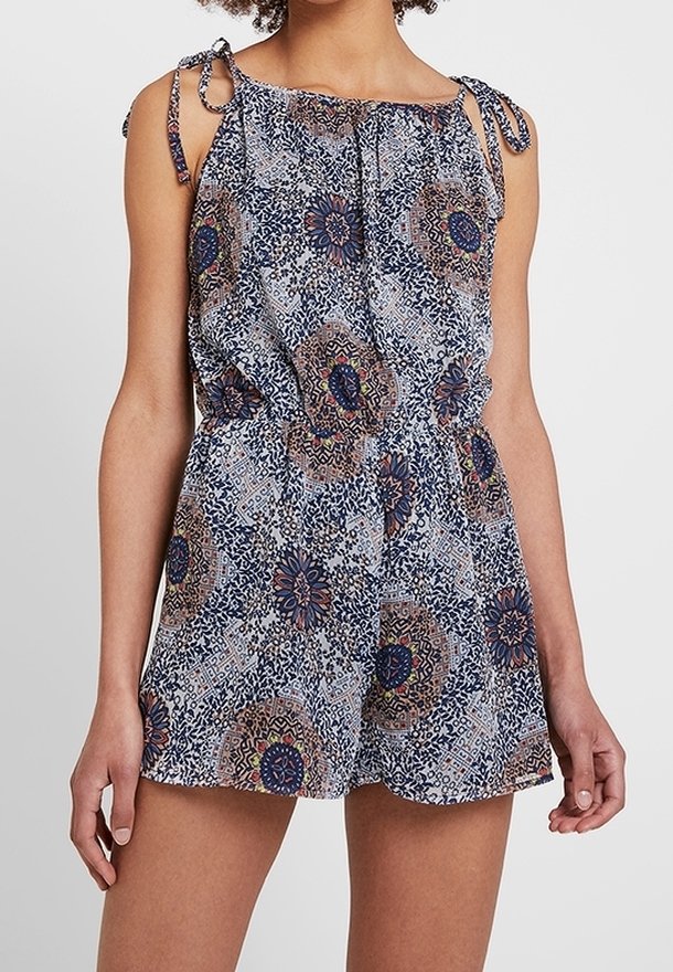 Femme portant une combinaison sans manches à motifs, avec des liens aux épaules et une taille élastique, présentant des motifs circulaires et floraux dans des tons bleus et bruns.