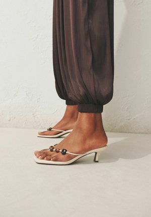 Jalad, mis kannavad pruune pärlitega kaunistatud rihmaga kõrgete kontsadega sandaale, kombineeritud lahtiste pruuni värvi elastsete sääristega pükstega heledal tekstuuriga seina taustal.