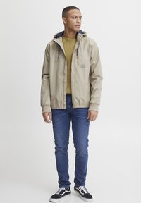 Beige vanntett jakke med blå hette, glidelås og logo-patch, kombinert med en grønn genser med rund hals og blå jeans. Sorte joggesko.