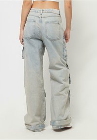 Persoană îmbrăcată cu pantaloni cargo din denim deschis, largi, cu tivuri răsucite și buzunare pe spate, stând pe un fundal simplu.