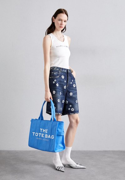 Borsa a tracolla blu in tela con testo bianco "THE TOTE BAG" e "MARC JACOBS." La modella indossa una canotta bianca e pantaloni corti di jeans blu con ricami floreali.