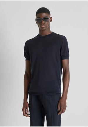 Giovane uomo con indosso una maglietta nera a maniche corte, pantaloni cargo neri e occhiali da sole neri, in piedi davanti a uno sfondo grigio chiaro uniforme.