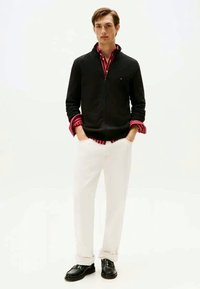 Black zip-up sweater over a red and white striped shirt, paired with white trousers and black shoes. Smooth fabric, casual fit, modern design.