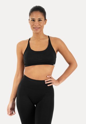 Ung kvinde iført sort sportsbh og leggings med høj talje, stående med den ene hånd på hoften, smilende til kameraet på hvid baggrund.