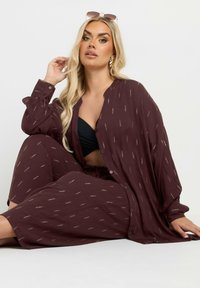 Camisa holgada de mangas largas en color marrón, con patrones sutiles; combinada con una blusa negra y pantalones a juego. Textura suave y diseño relajado.