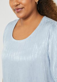 Helles blaues Blusenshirt mit Rundhalsausschnitt, strukturiertem Stoff mit vertikalen Streifen und kurzen Puffärmel. Glattes Material mit subtiler Glanz.