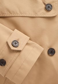 Beige trenchcoat i närbild med mjuk textur, två svarta knappar med blomsterdesign och skarpa kragdetaljer.