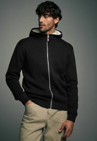 Mustmust musta jakks, millel on pehme valge sisemine vooder, soonikkoes varrukad ja äär. Mudel kannab seda khaki pükstega. Lihtne, puhas disain.