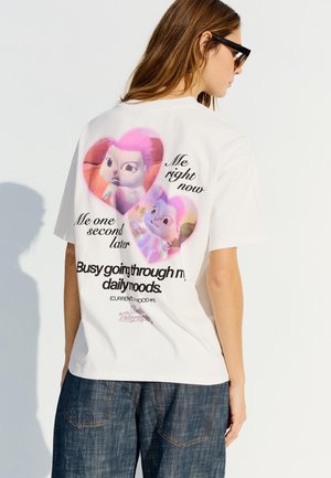 Woman wearing white T-shirt with two animated cat faces inside hearts and text about daily moods on the back, paired with blue jeans.