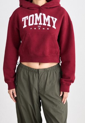 Osoba ubrana w krótki bordowy kaptur z logo "TOMMY JEANS" i luźne oliwkowozielone spodnie, stojąca na tle jasnego, jednolitego tła.