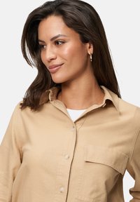 Beige overhemd met knopen, een kraag, lange mouwen en een borstzak. De stof heeft een gladde textuur en bevat subtiele stikdetails.