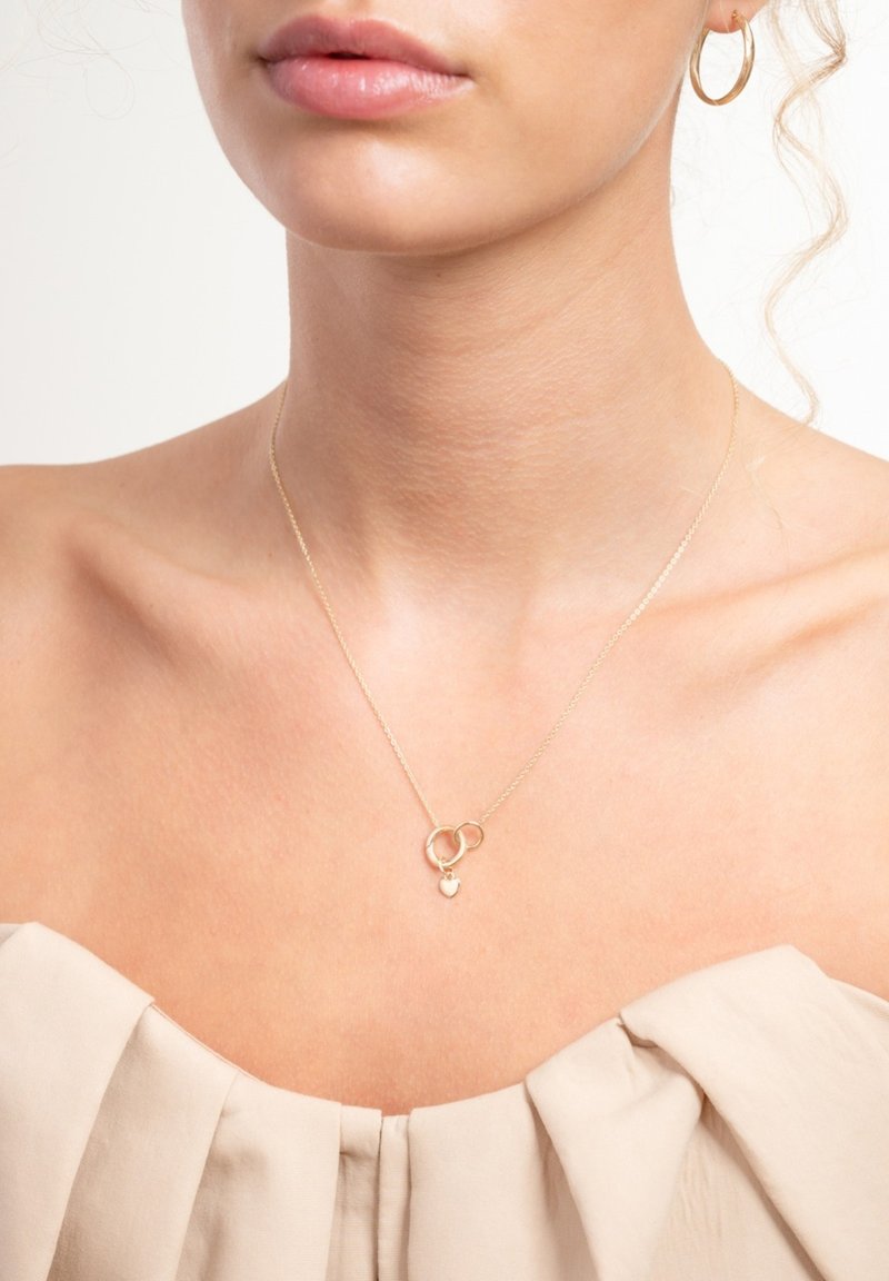 Gouden halsketting met twee in elkaar grijpende ringen en een klein hartvormig hangertje, getoond op een blote hals. Zachte textuur en delicaat kettinkje.