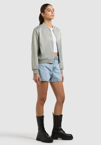 Femme portant une veste bombardier argentée, un crop top blanc, un short en jean clair et des bottes de combat noires à lacets, debout devant un fond gris.