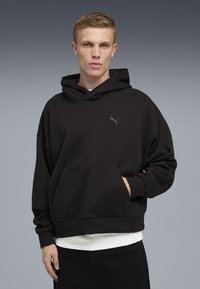 Ung mand med kort blondt hår, iført en sort Puma-hoodie og hvid skjorte, stående mod en ensfarvet grå baggrund.