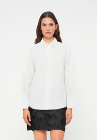 Camicia bianca con colletto impreziosito, abbinata a una gonna nera ricamata con motivi floreali e orlo grezzo.