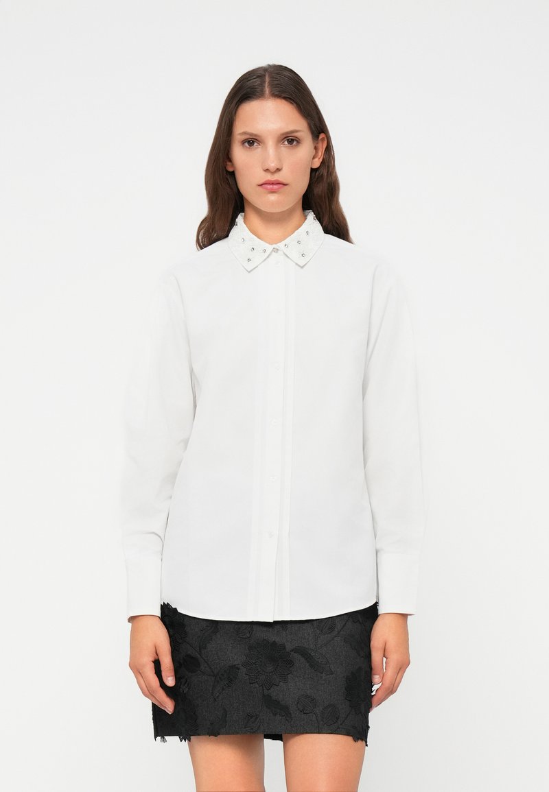 Camicia bianca con colletto impreziosito, abbinata a una gonna nera ricamata con motivi floreali e orlo grezzo.