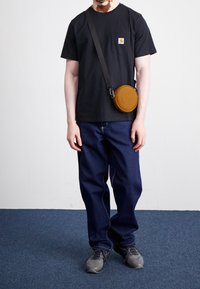 Černé tričko s krátkým rukávem a kapsou; modré široké džíny; kulatá béžová kabelka přes rameno; černé sportovní boty. Hladký pozadí.