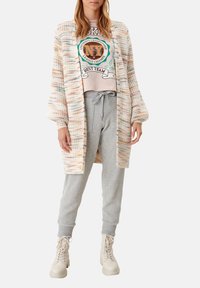 Mehrfarbiger Strickcardigan mit langen Ärmeln, offenem Vorderteil und strukturiertem Muster. Getragen über einem Grafik-T-Shirt und grauen Jogginghosen mit Schnürung. Beigefarbene Chunky-Boots.