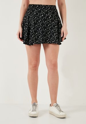 Žena u crnoj mini suknji s cvjetnim uzorkom i bijelim tenisicama, stoji ispred jednostavne svijetle pozadine.