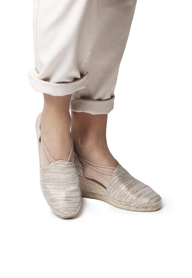 Beige espadrilles wedges dalam kain dengan tekstur garis-garis, dilengkapi sol fleksibel dan tali pergelangan kaki. Celana berwarna terang yang digulung terlihat.