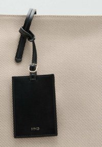 Sac en toile beige avec un relief diagonal, doté d'une étiquette en cuir noir accrochée par une sangle et d'un détail de marquage doré.