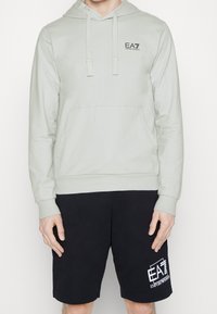 Uomo che indossa una felpa EA7 Emporio Armani grigia chiara con lacci e pantaloni corti neri con logo EA7 su uno sfondo bianco.