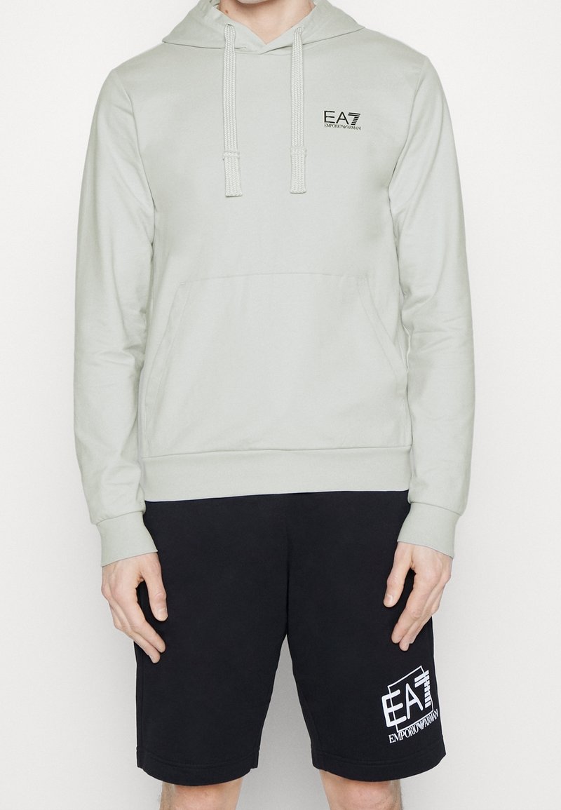 Uomo che indossa una felpa EA7 Emporio Armani grigia chiara con lacci e pantaloni corti neri con logo EA7 su uno sfondo bianco.