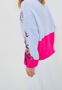 Sweter z dzianiny w jasnoniebieskich i jaskrawo różowych częściach, z kwiatową haftowaną dekoracją na rękawach i prążkowanymi mankietami dla tekstury.