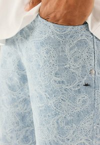 Mano en el bolsillo descansando sobre unos jeans de mezclilla azul claro con bordado de paisley blanco y pequeños botones negros y plateados.