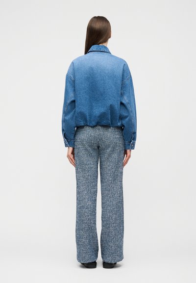 Femme debout de dos, portant une veste en jean bleue et un pantalon gris texturé taille haute avec des chaussures noires.