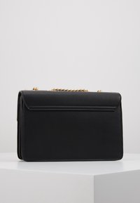 Borsa a mano nera in pelle rettangolare con tracolla in catena dorata, poggiata su una superficie bianca contro uno sfondo grigio chiaro uniforme.
