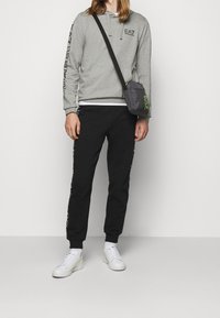 Szara bluza z kapturem z logo, czarne spodnie dresowe z nadrukiem z boku, białe sneakersy oraz szara torba na ramię. Prosty, sportowy design i komfortowe dopasowanie.