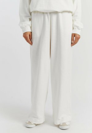 Weiße, weit geschnittene Hosen aus weichem Stoff, mit elastischem Bund und glatter Textur. Kombiniert mit hellfarbigen Schuhen.