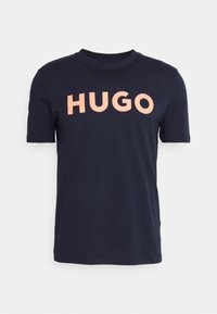 Navy sininen puuvillapaita, jossa on pyöreä kaula-aukko ja lyhyet hihat. Edessä on iso persikan värinen teksti, jossa lukee "HUGO".