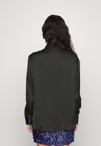 Černá hedvábná košile na knoflíky s hladkou texturou, dlouhými rukávy a zakřiveným lemem, nošena přes sukní s flitry v mnoha barvách.