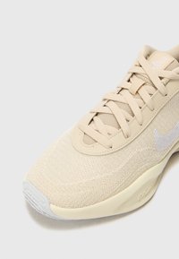 Chaussure de course Nike beige à tige en maille, lacets en tissu, logo swoosh blanc, et semelle amortie, présentée en angle pour montrer les détails avant et latéraux.