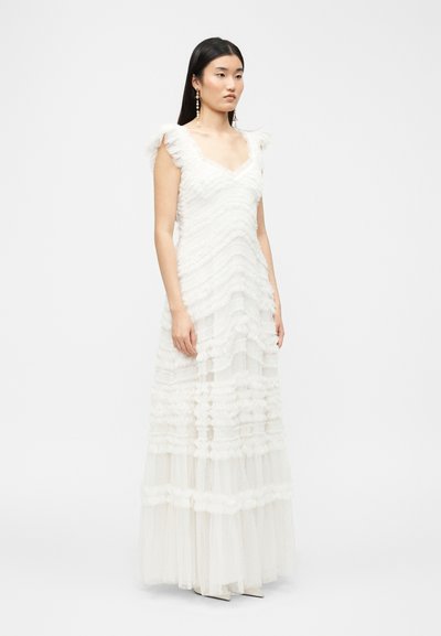 Γυναίκα με μακριά σκουρόχρωμα μαλλιά που φορεί ένα λευκό μακρύ φόρεμα με βολάν χωρίς μανίκια και μακριά κρεμαστά σκουλαρίκια, στέκεται μπροστά σε απλό φόντο.