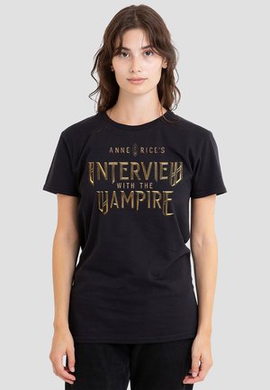 Young woman with brown hair wearing a black T-shirt with gold text "Anne Rice's Interview with the Vampire" on a light gray background.