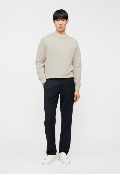 Μπεζ φούτερ με στρογγυλή λαιμόκοψη, συνδυασμένο με σκούρο παντελόνι. Το μοντέλο φοράει λευκά αθλητικά παπούτσια. Ομαλή υφή και άνετη εφαρμογή.