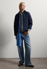 Giacca imbottita blu navy, camicia azzurro chiaro, jeans larghi in denim chiaro, abbinati a una sciarpa blu e scarpe marroni scure. Tessuti morbidi e vestibilità rilassata.