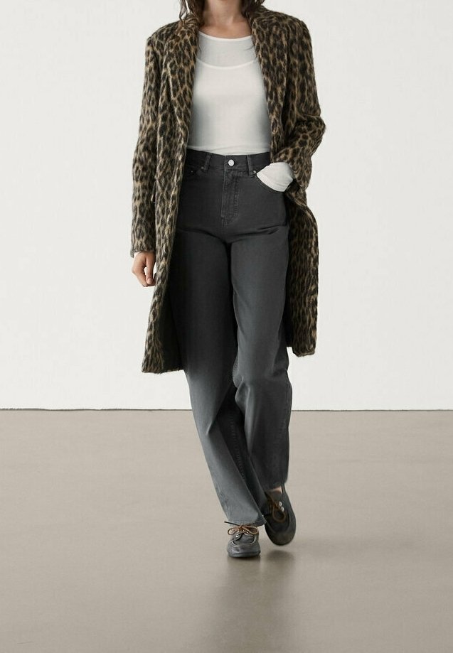 Personne portant un long manteau à imprimé léopard, un haut blanc, un pantalon taille haute gris foncé et des chaussures grises à lacets marchant vers l'avant.