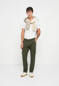 Grüne Cargo-Hosen mit Seitentaschen, kombiniert mit einem schlichten weißen T-Shirt und einem hellbeigen Pullover, der über die Schultern drapiert ist. Cremefarbene Sneaker sind ebenfalls zu sehen.