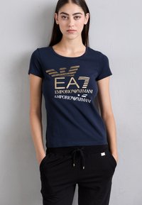 Tamsiai mėlynas marškinėlis su auksine grafine užrašu "EA7 Emporio Armani", turintis prigludusį dizainą. Trumpi rankoviai, minkšta medžiaga, derinamas su juodais džinsais.